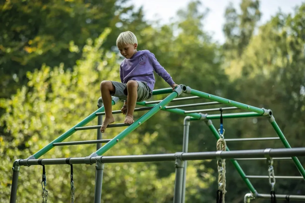 Junge sitzt oben auf einem grünen Klettergerüst und schaut über den Garten