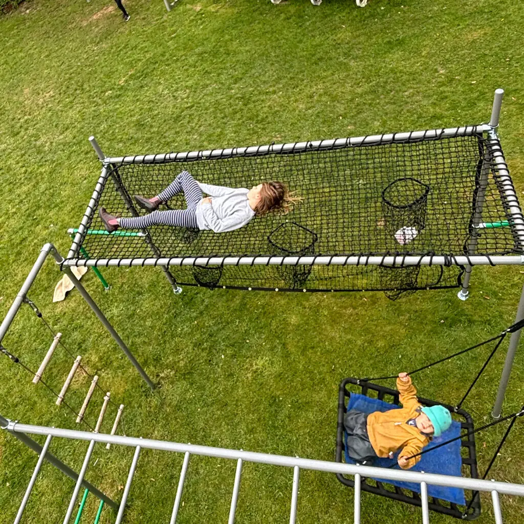 Kind liegt entspannt im FOURACTION Liegenetz am Klettergerüst, daneben Kletterleiter und Schaukelsitz im Garten