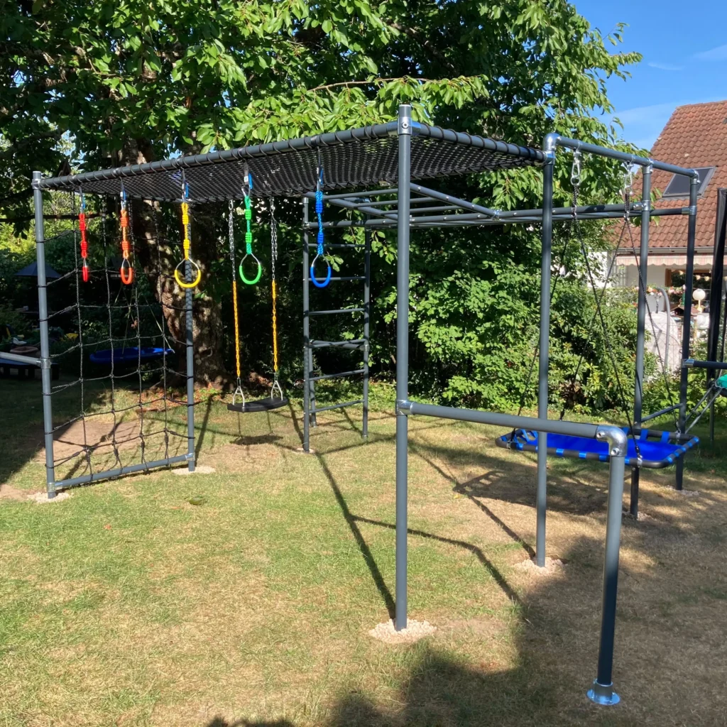 Little Action Klettergerüst BOLLE im Garten mit Liegenetz, Turnringen in Rot, Gelb, Grün und Blau, Strickleiter, Schaukel und blauer Plattformschaukel, umgeben von Bäumen und Rasen