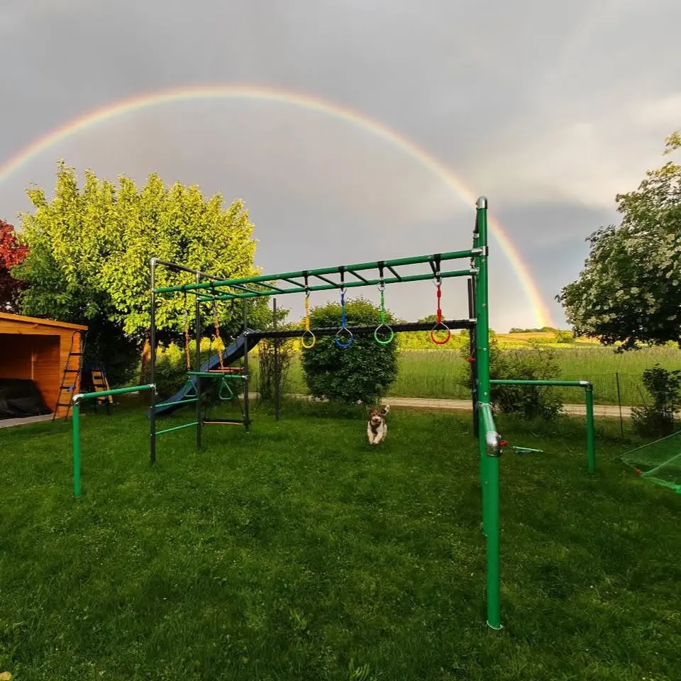 Little Action Klettergerüst BOLLE Grün im Garten mit bunten Turnringen, Trapez und Rutsche – im Hintergrund ein Regenbogen