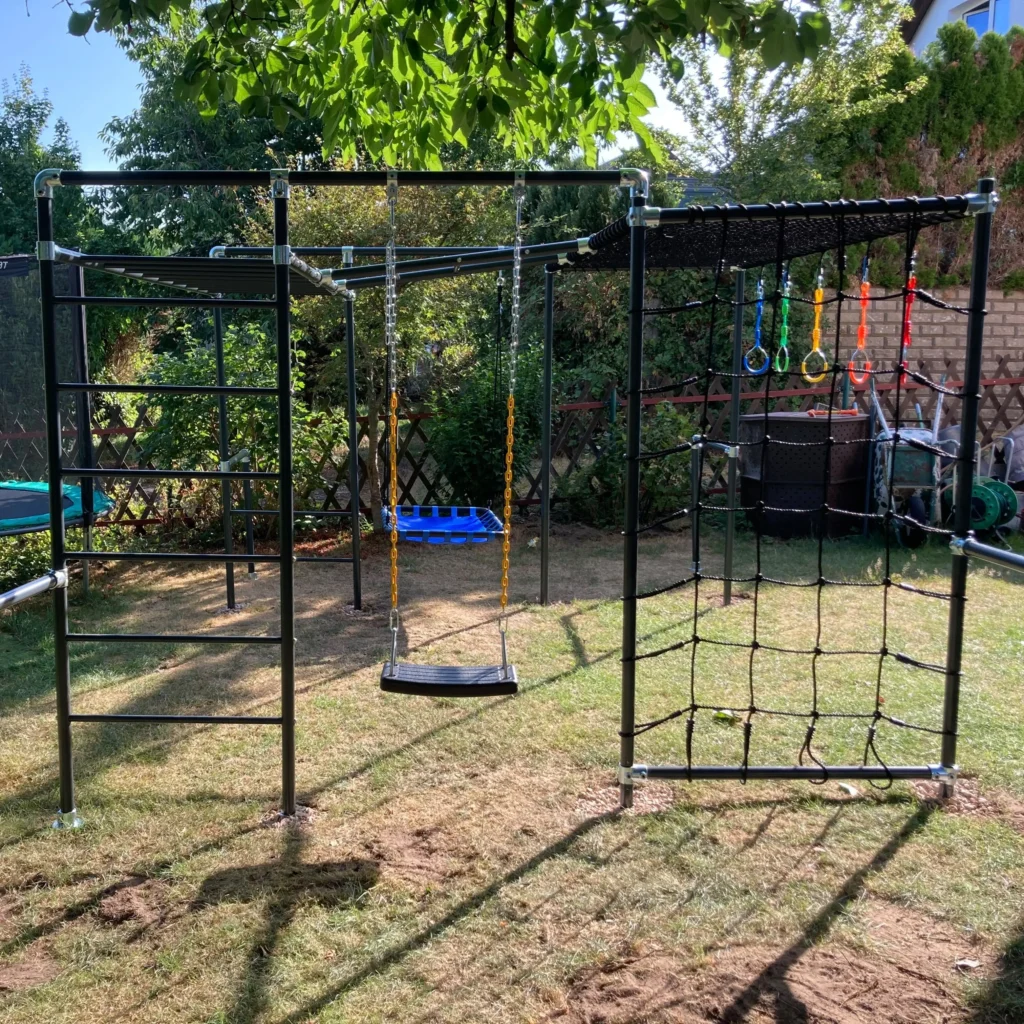 Little Action Klettergerüst BOLLE im Garten mit Sprossenwand, Schaukel mit blauem Plattformsitz und Turnringen in Blau, Grün, Gelb und Rot vor einem senkrechten Kletternetz, im Hintergrund Trampolin und Gartenzaun